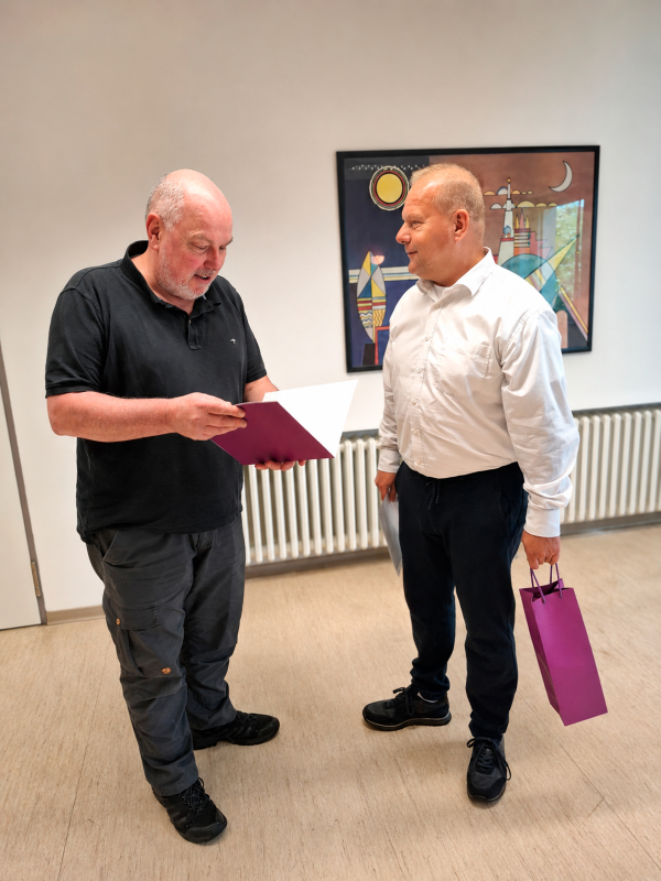 Zwei Männer stehen beieinander. Der Mann links, Michael Kostros, schaut auf eine Mappe. Der Mann rechts, Gerd Schwesig, lächelt und hält eine Geschenkpackung in der Hand.