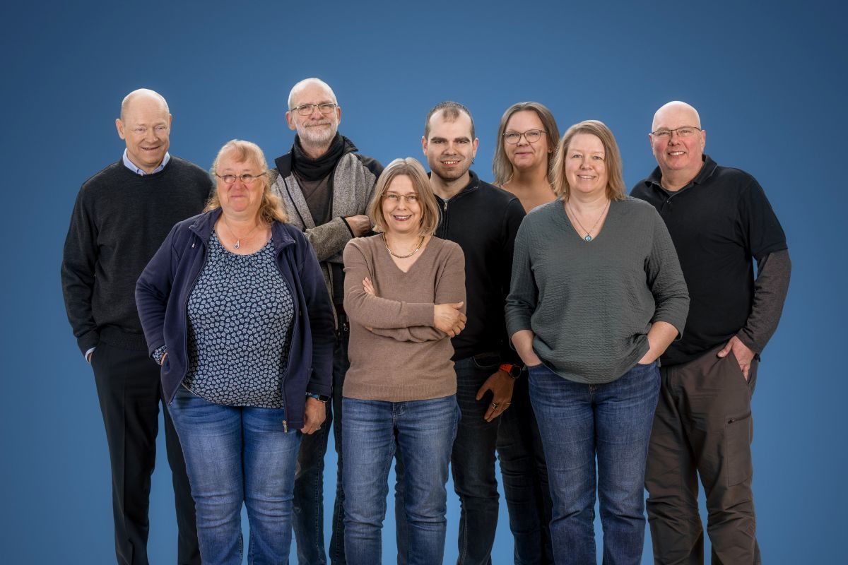 Vier Männer und vier Frauen stehen als Gruppe beieinander. Der Hintergrund ist blau. Sie lächeln in die Kamera. Die meisten von ihnen haben die Hände in den Hosentaschen.